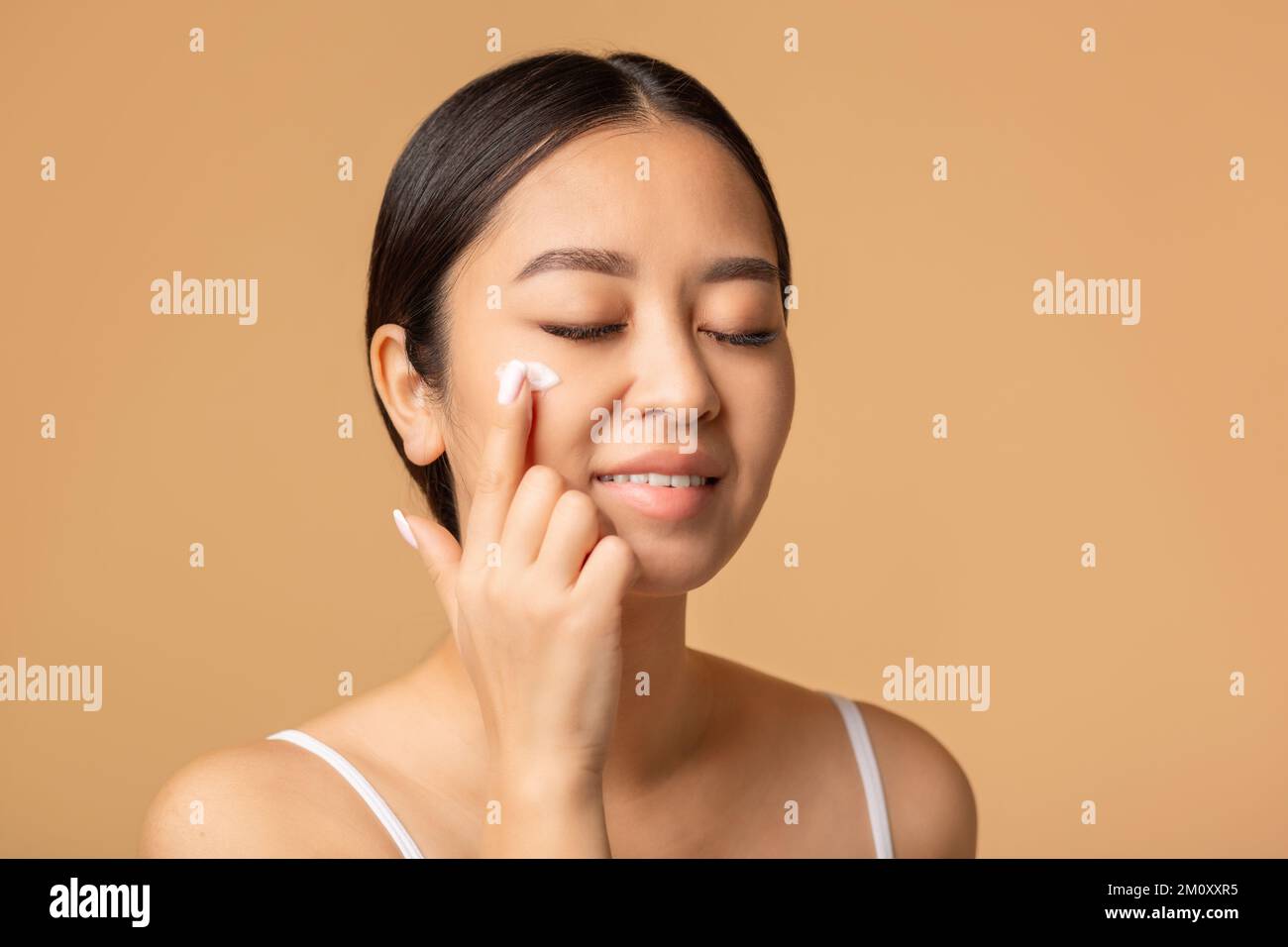 woman applying a anti age or moisturizer face cream on her face, smiling sweetly standing on beige background Stock Photo