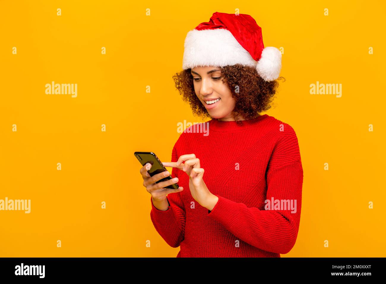 Young woman in santa claus hat holding mobile phone, reading holiday message, congratulates friends happy new year Stock Photo