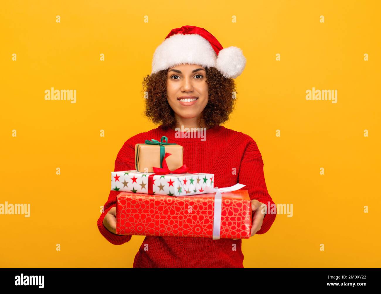 Happy woman holds in hands stack of christmas gifts. female in santa claus hat on orange background, smiling. Xmas Stock Photo