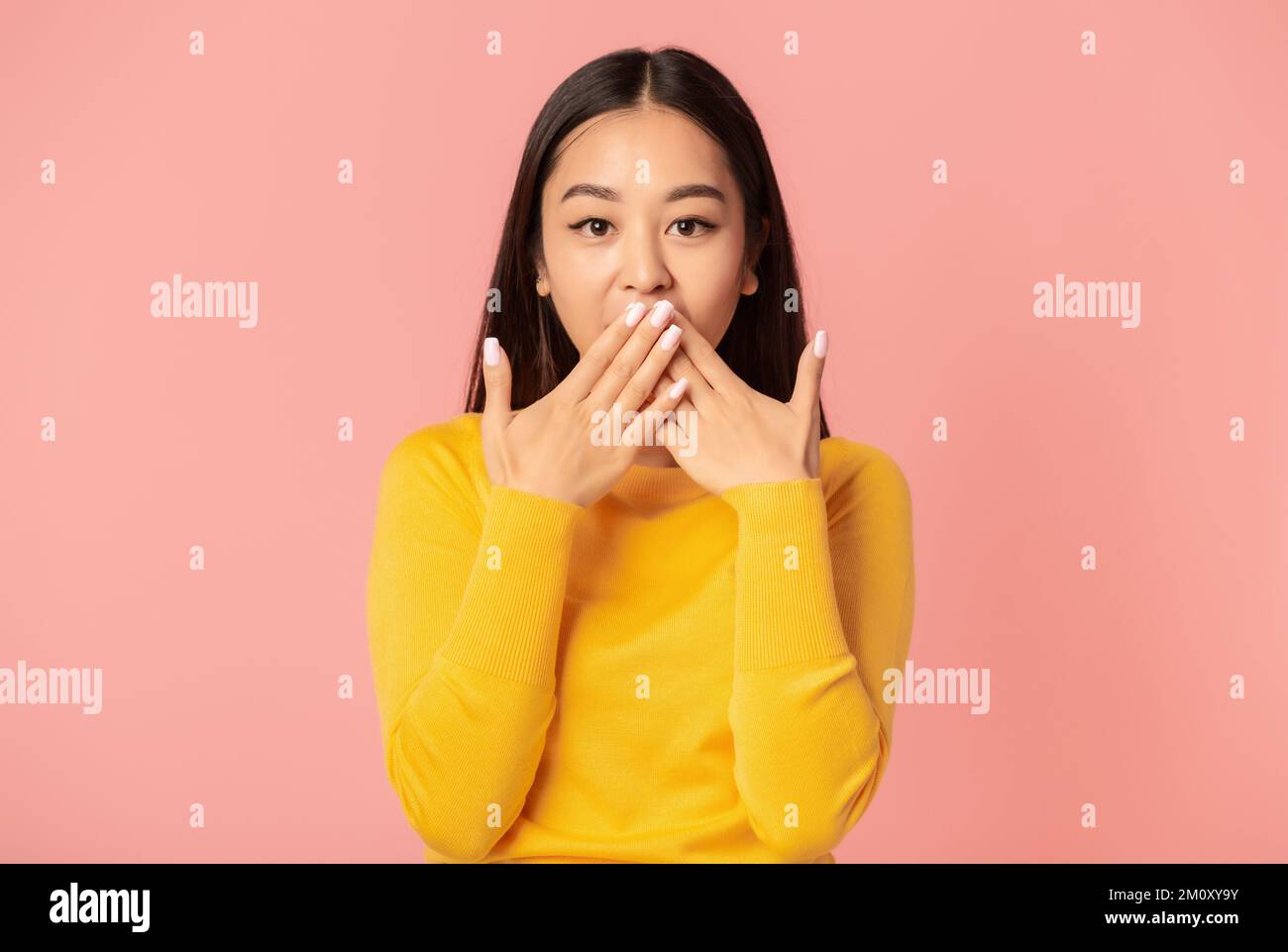 Surprised woman covering her mouth with hands while standing on pink background looking at the camera Stock Photo