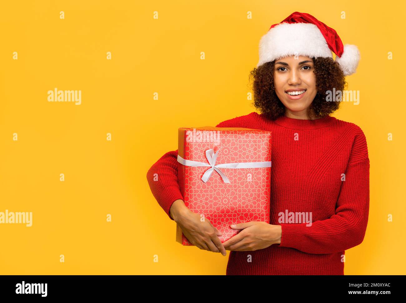 Happy African American woman holds in hands christmas gift Female in santa claus hat on orange background, smiling. Xmas Stock Photo