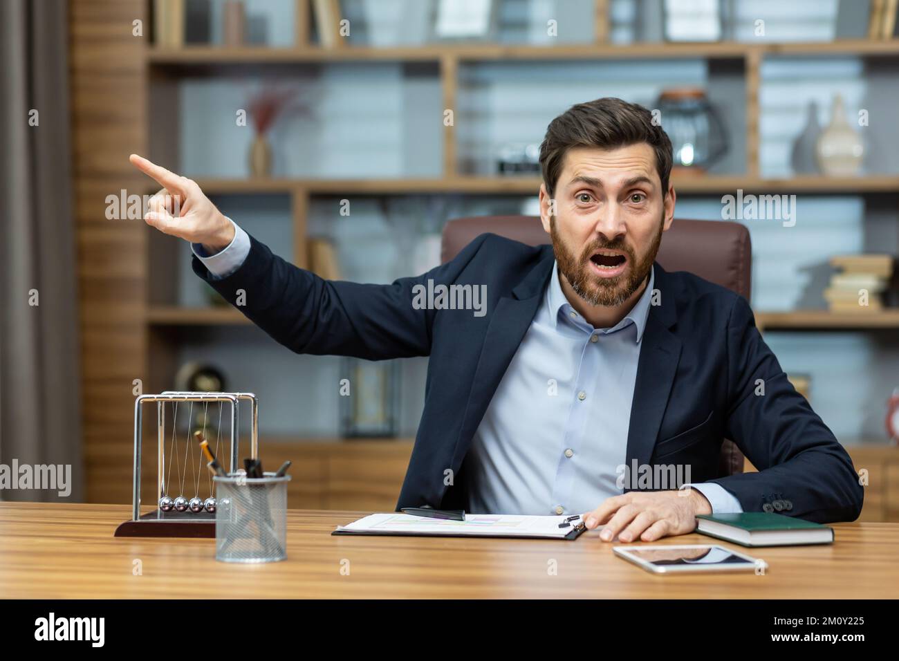 Angry boss shouts at the camera to the employee points his finger to the side dismisses the subordinate, a businessman in a business suit sits at a table in a modern office. Stock Photo