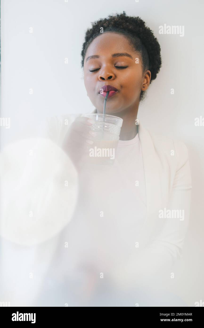 Beautiful african girl wearing white jacket outfit holding an ice coffee sitting in a cafe shop against the white wall. A pleasant pastime at Stock Photo