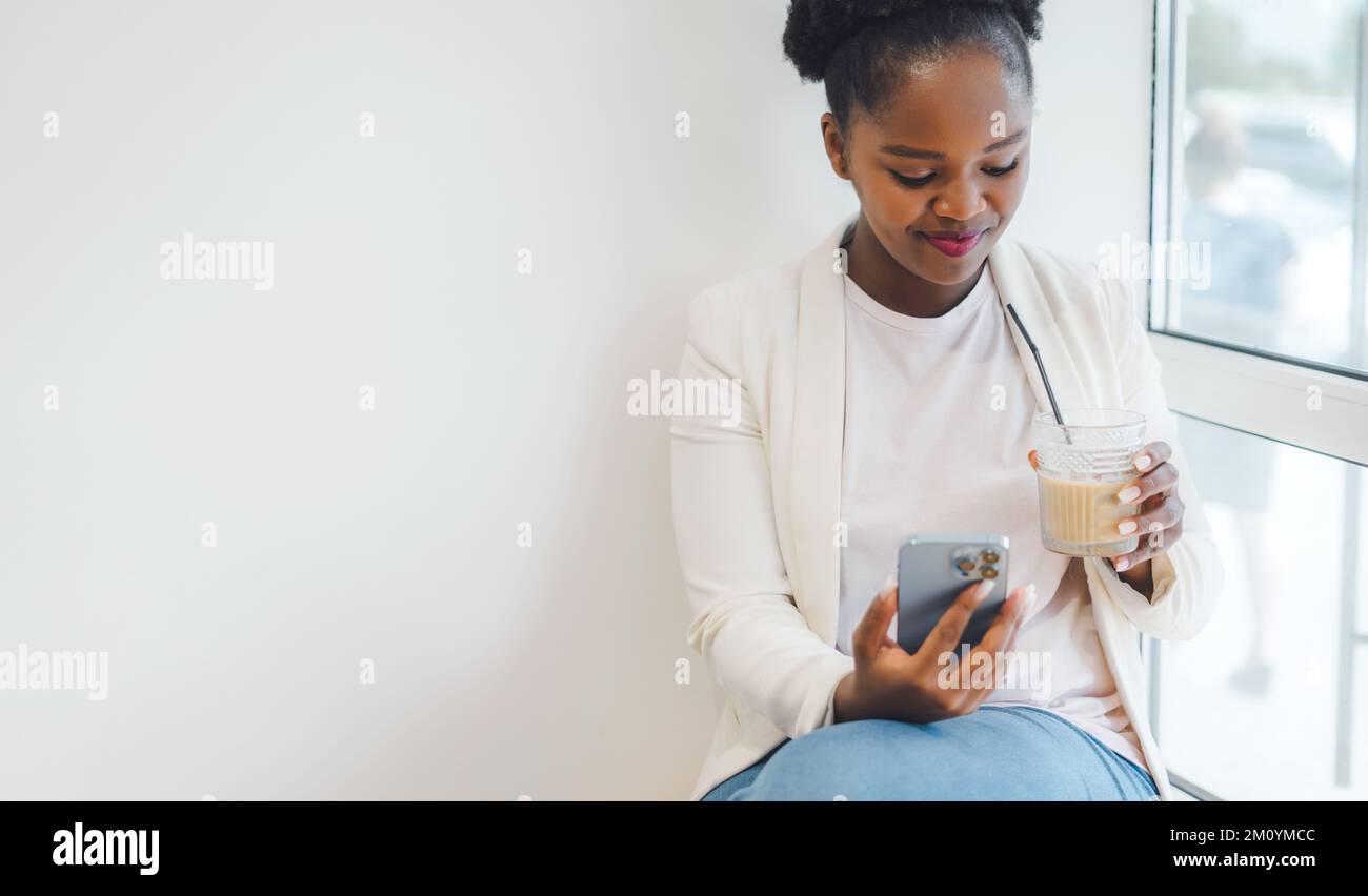 A business african woman sitting at a table in a cafe, drinking ice coffee and looking through her phone, wearing a white jacket. Coffee break in a Stock Photo