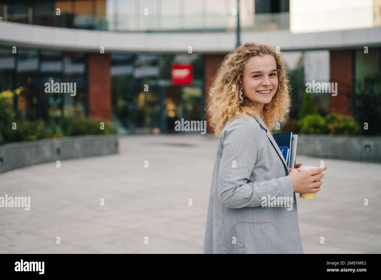 Stylish city girl with curly hair holding laptop in her hands, walking on street and drinking takeaway coffee, going to university. Girl student Stock Photo