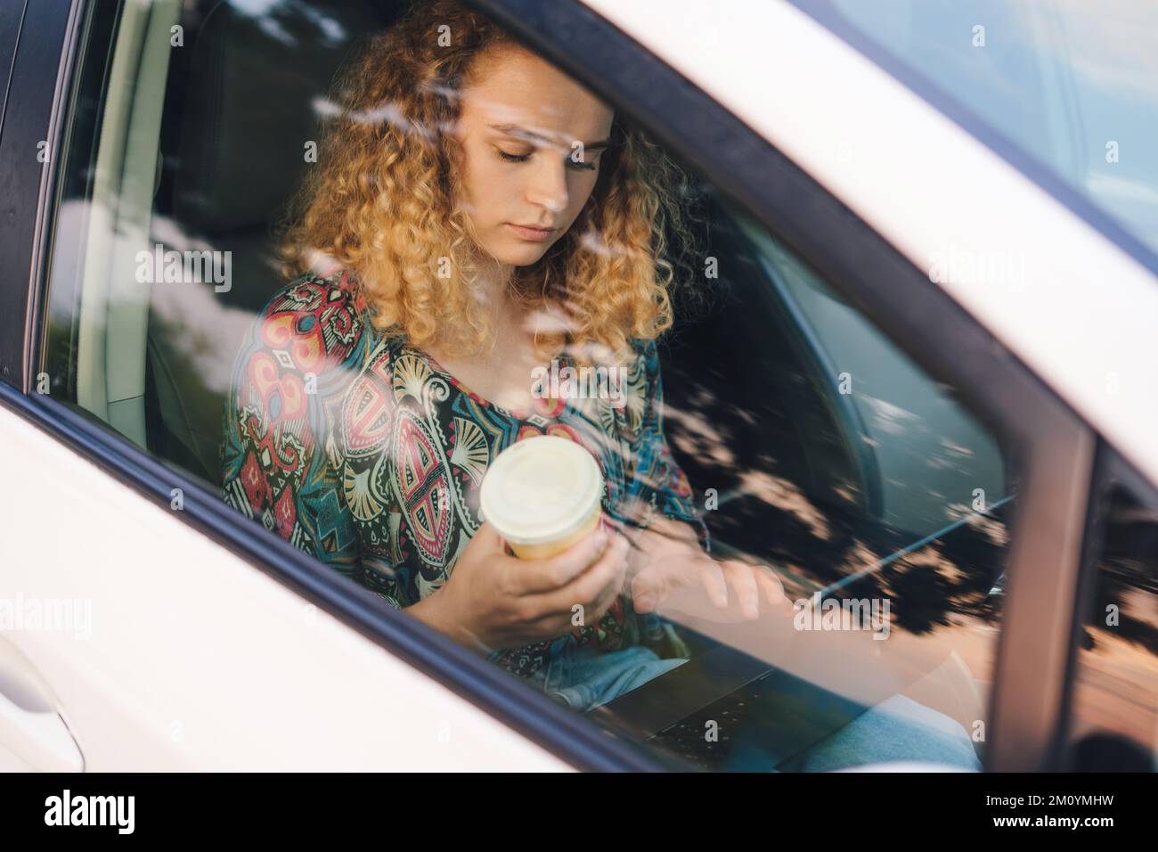 Young business woman sitting in the car drinking coffee and texting on a laptop. View from car window. Business people lifestyle. Stock Photo