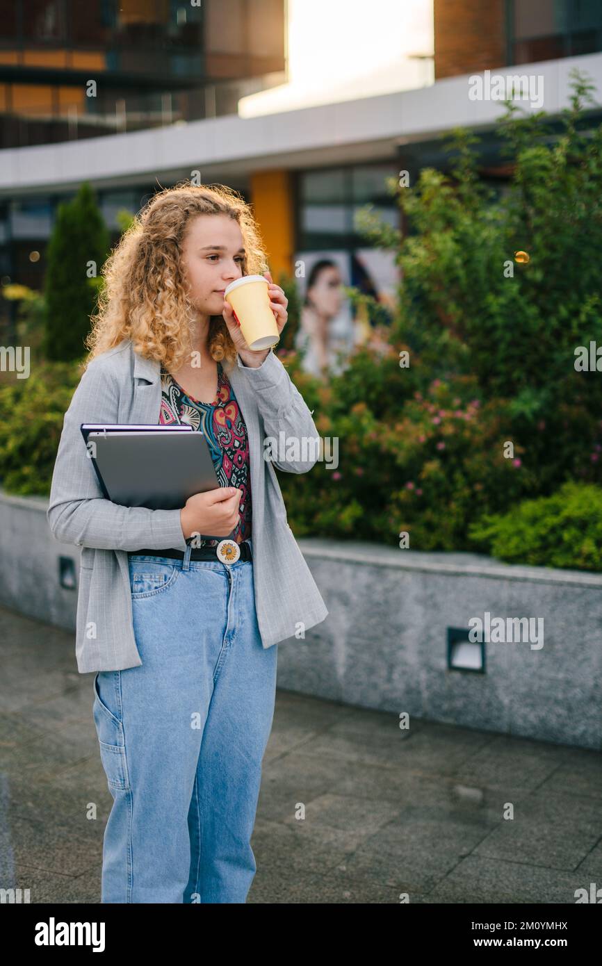 Curly-haired caucasian girl student having morning coffee while going to university, holding her laptop in hands. Lifestyle, college and people Stock Photo