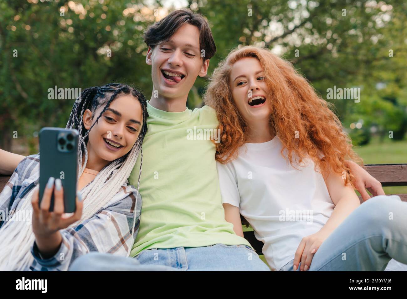 Three caucasian smiling friends at the park sitting on a bench and taking selfies using a smart phone. Summer vacation fun. Having fun. Stock Photo