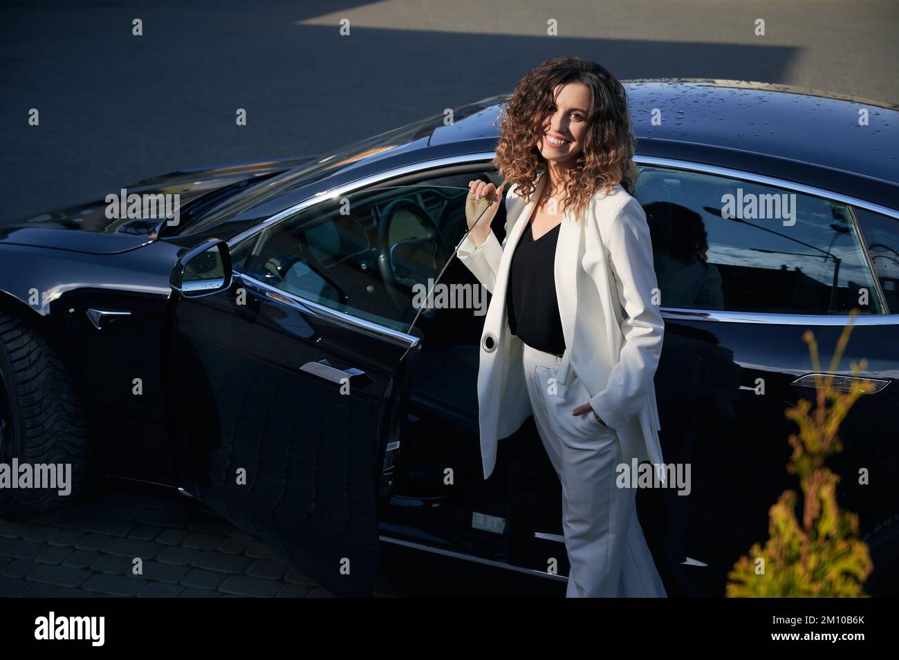 Attractive businesswoman looking at camera and smiling while standing by electric vehicle on the street. Cheerful young woman wearing stylish white suit while posing near electric automobile outdoors. Stock Photo