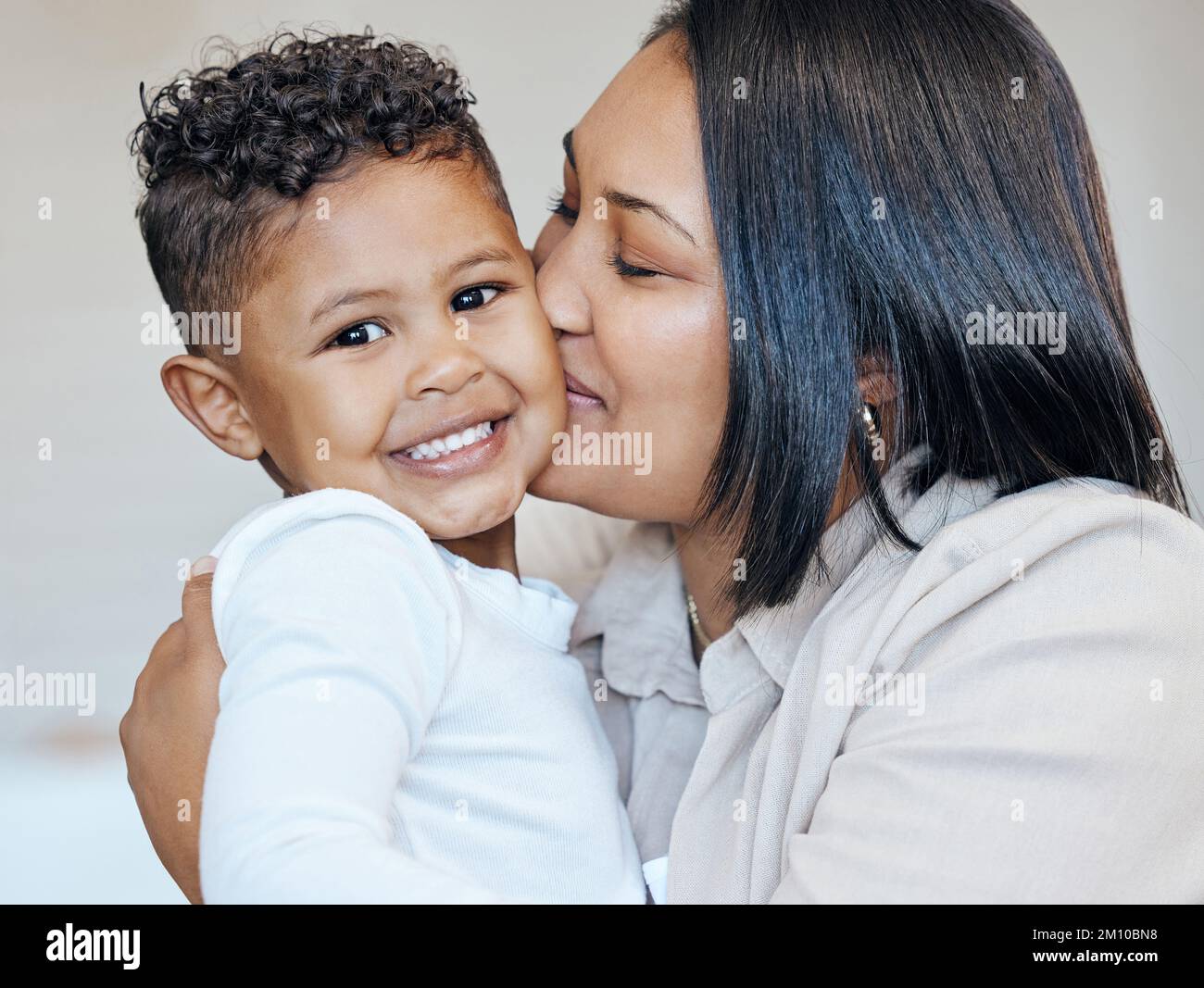 Mixed race woman kissing her adorable little son on the cheek while bonding together at home. Small hispanic boy smiling and looking happy to be Stock Photo