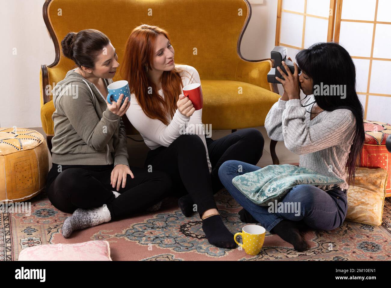 Young female friends having fun indoors taking pictures of each other with vintage polaroid camera. Multiethnic group of millennials Stock Photo