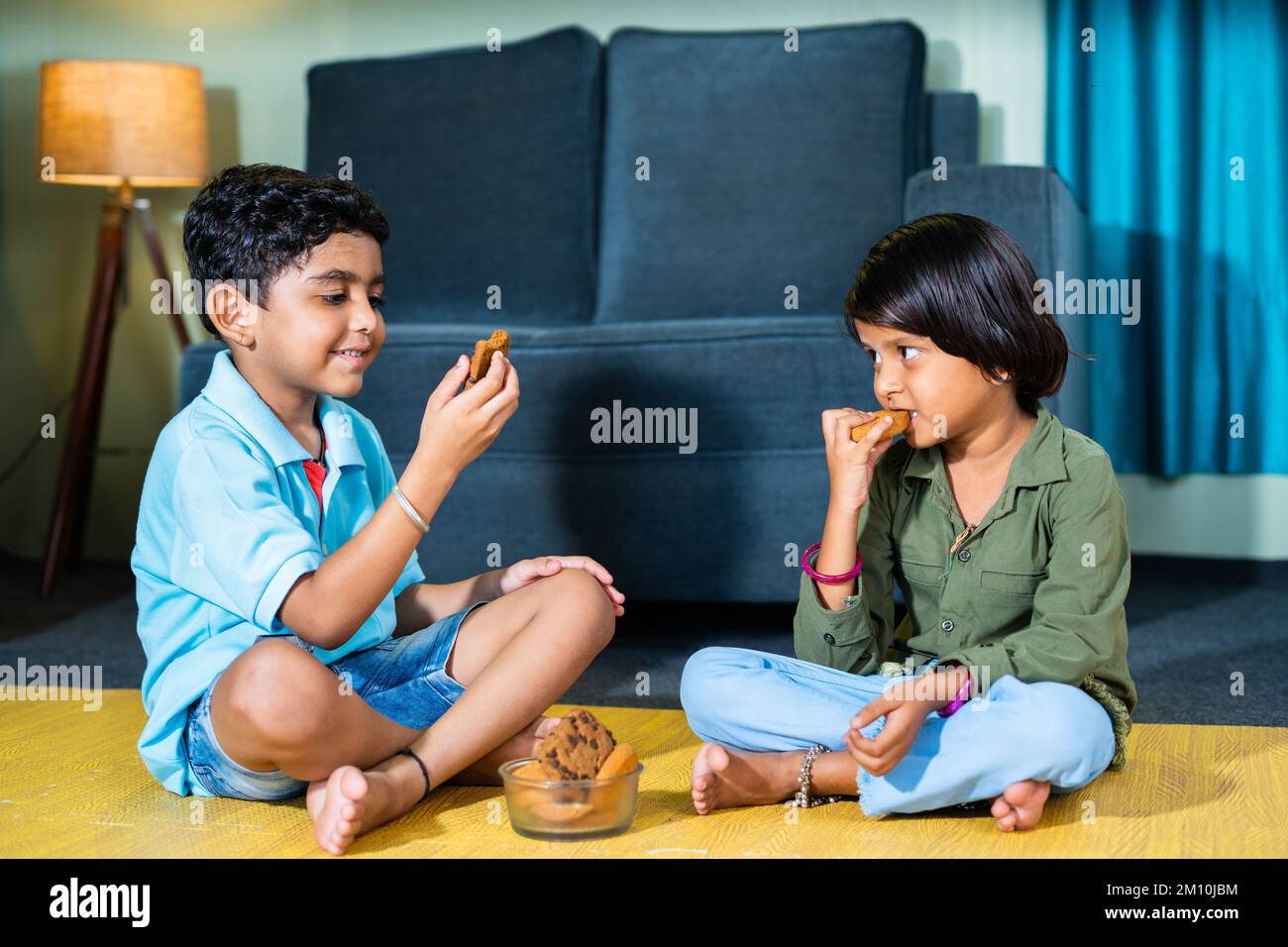 Young sibling kids having snacks or biscuit while sitting on floor at home by looking around after school - concepts of healthy eating togetherness Stock Photo