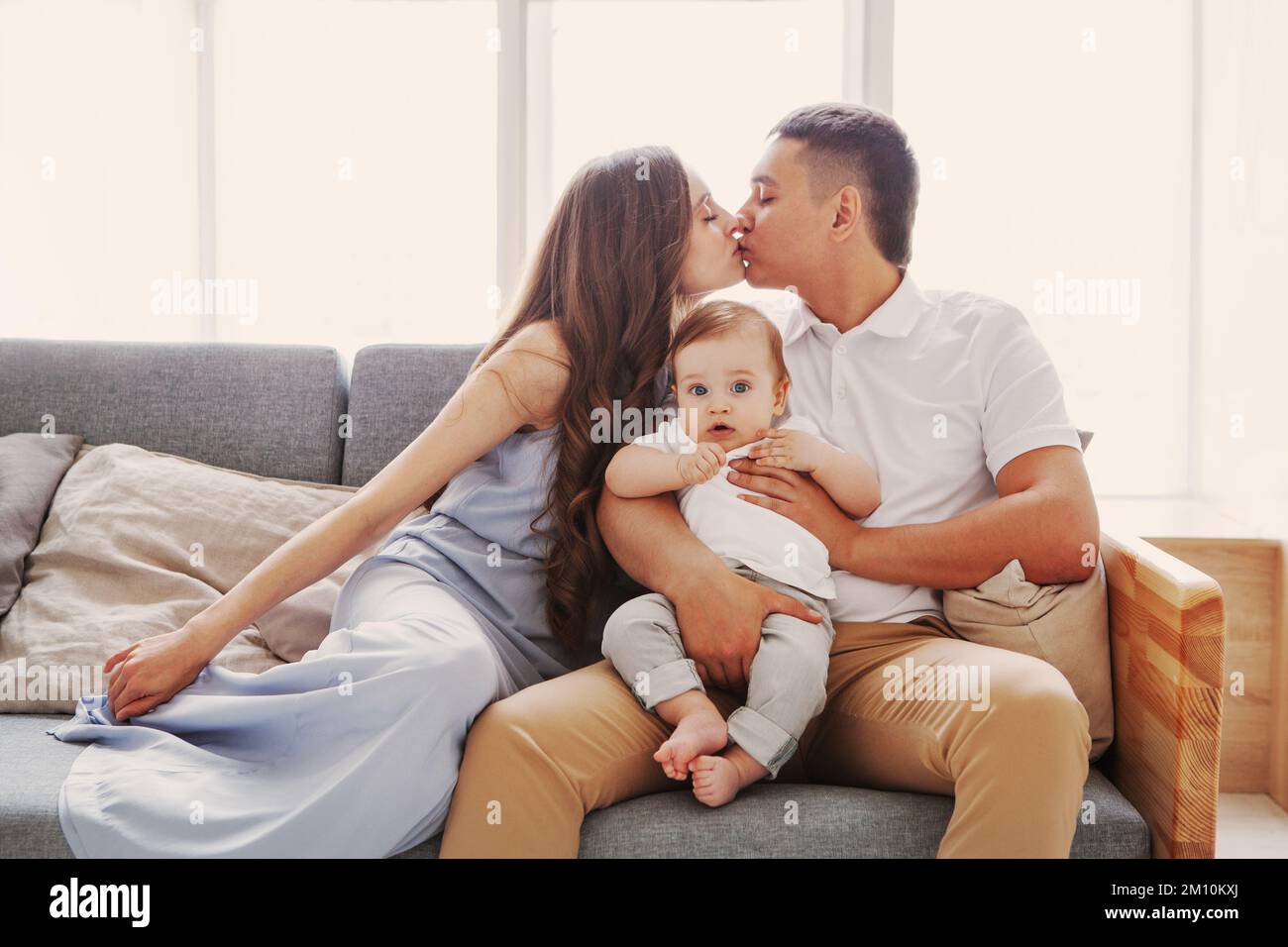 Mom and Dad kissing while the child looking at camera. Happy parents sitting home on couch in front of the window. Family relationship and love Stock Photo