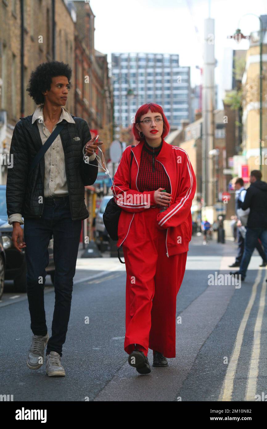 Cool street style man with afro hairstyle, wearing jeans, jacket and white trainers walks with woman with red hair and red tracksuit on London street Stock Photo