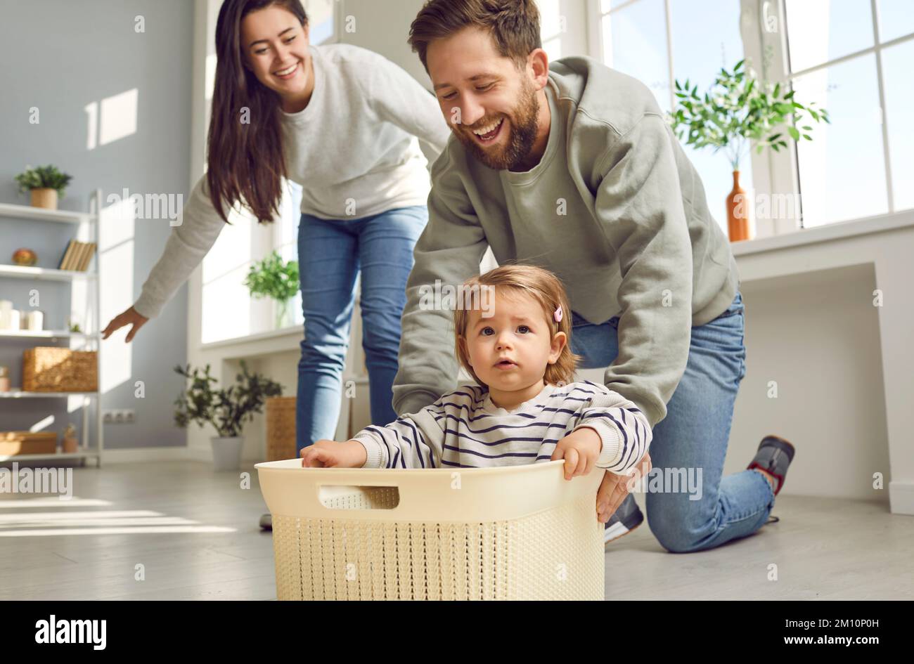 Cheerful young parents are playing with their little daughter, carrying her around house in box. Stock Photo