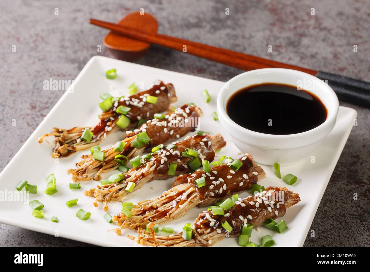 Delicious fried beef roll with enoki mushrooms with teriyaki sauce, green onions and sesame close-up in a plate on the table. Horizontal Stock Photo