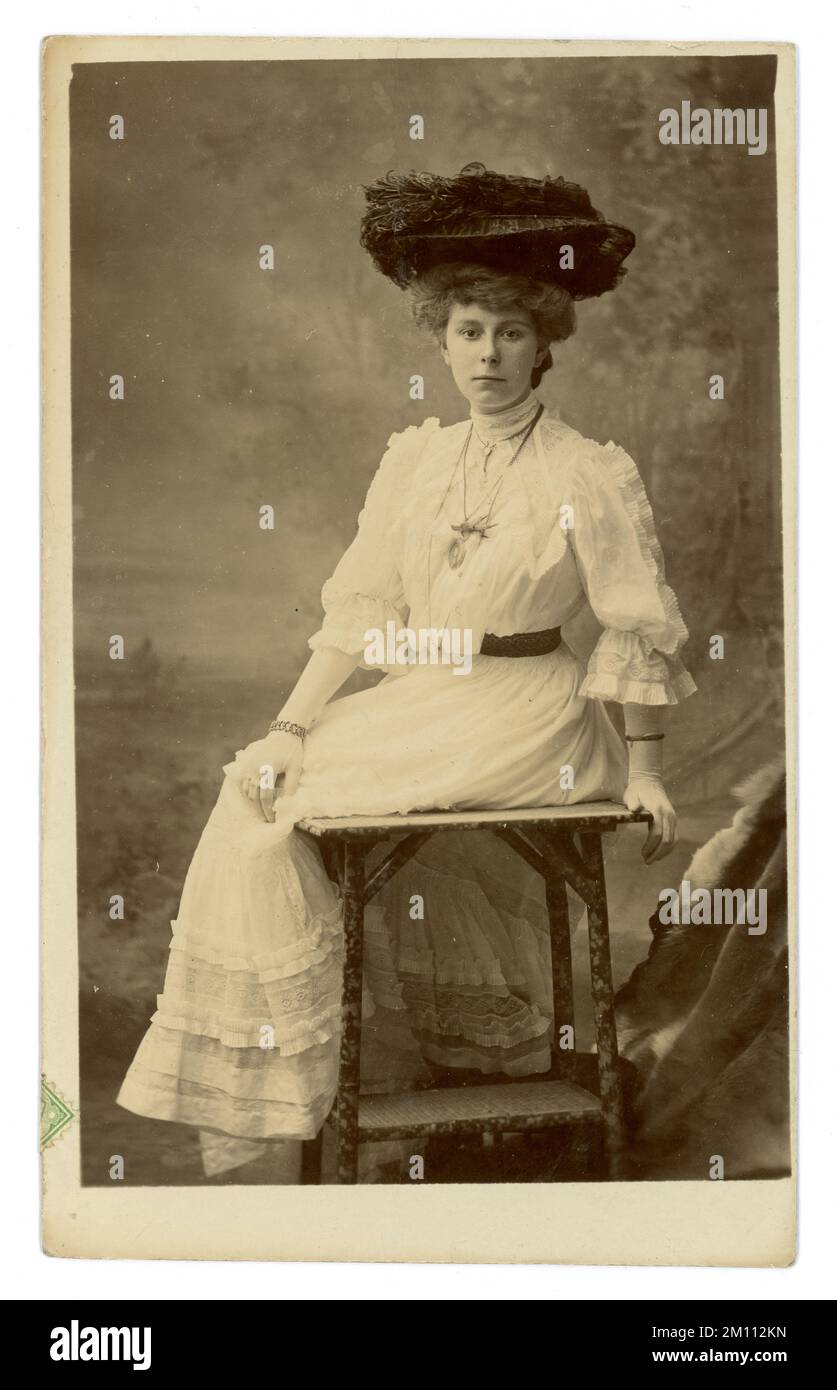 Original Edwardian era postcard of beautiful lady, wearing a large elaborate hat  trimmed with ostrich feathers, wearing a white summer dress, flowing sleeves, gloves, a bamboo table prop typical of the furniture of this period. Edwardian lady. Edwardian woman. Dated / posted 20 November 1906, Sheffield, England, U.K. Stock Photo