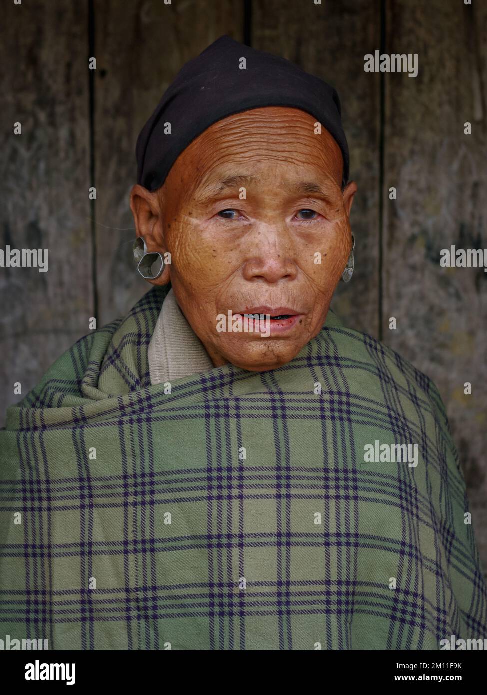 Mon, Nagaland, India - 03 03 2009 : Frontal portrait of beautiful old Naga tribe woman wearing green plaid and black scarf on dark wooden background Stock Photo