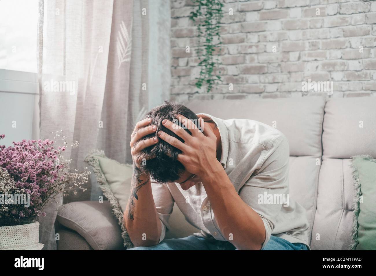 One young sick man sitting on the sofa feeling bad with pain and headache. Teenager with a tattoo on his arm touching his face unwell. Stock Photo