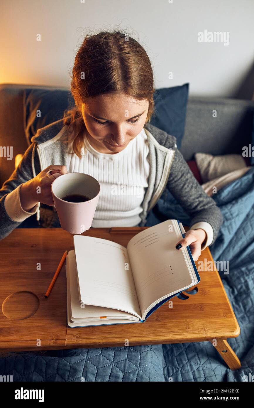 Student learning at home. Young woman making notes, reading and learning from notepad. Girl writing journal sitting in bed at home during quarantine Stock Photo