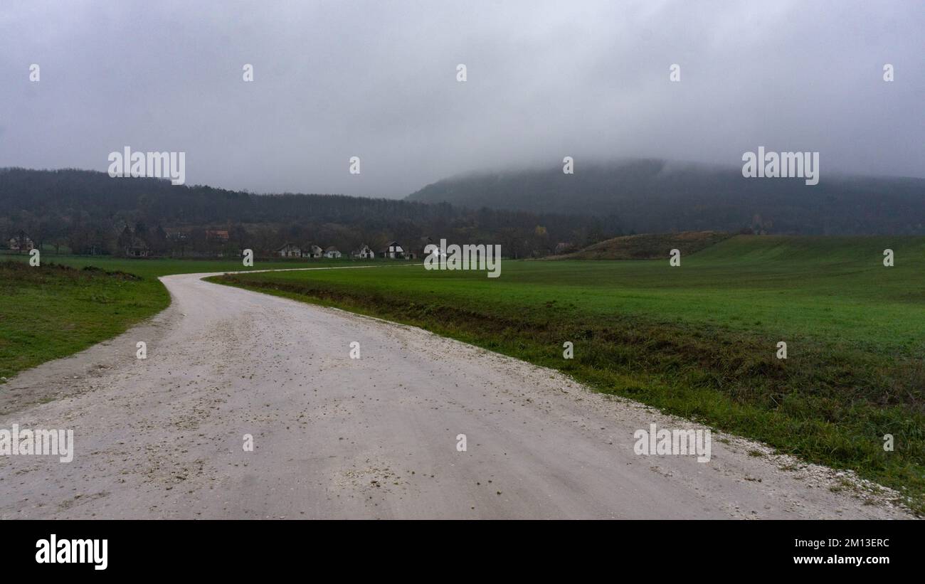 Misty dirt road goes into the little village and the hills over a green pasture in dark cloudy weather in Pilis, Hungary Stock Photo