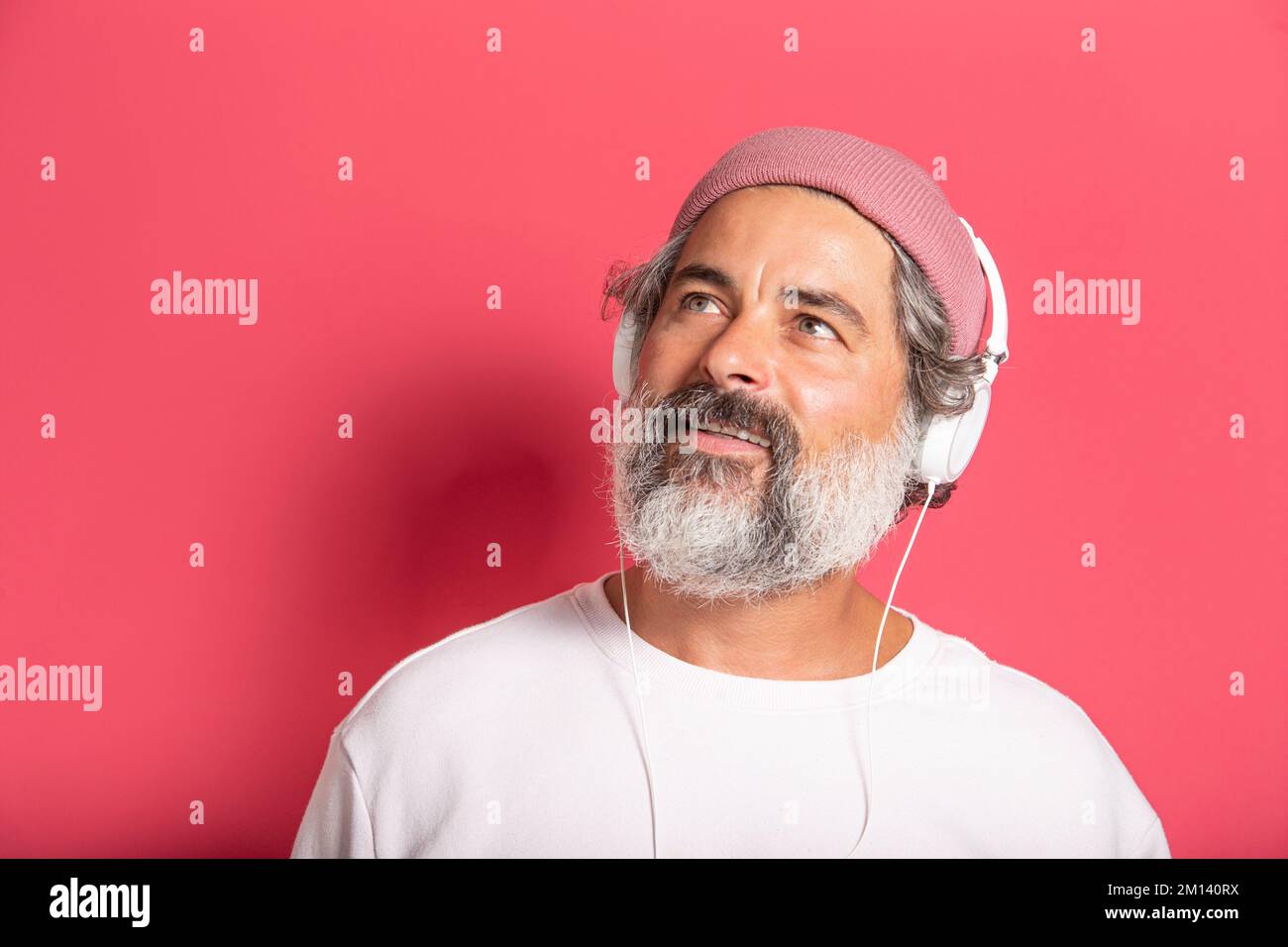 A Caucasian Cheerful gray-haired bearded man in casual pink hat and pullover listening to music with headphones isolated on fuchsia wall background Stock Photo