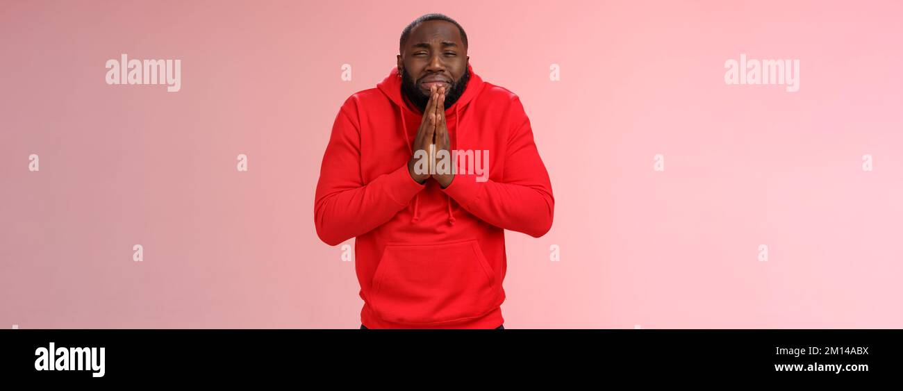 Miserable african american man beard in red hoodie stooping make supplicating upset grimace grying sobbing asking help say please hold hands pray Stock Photo
