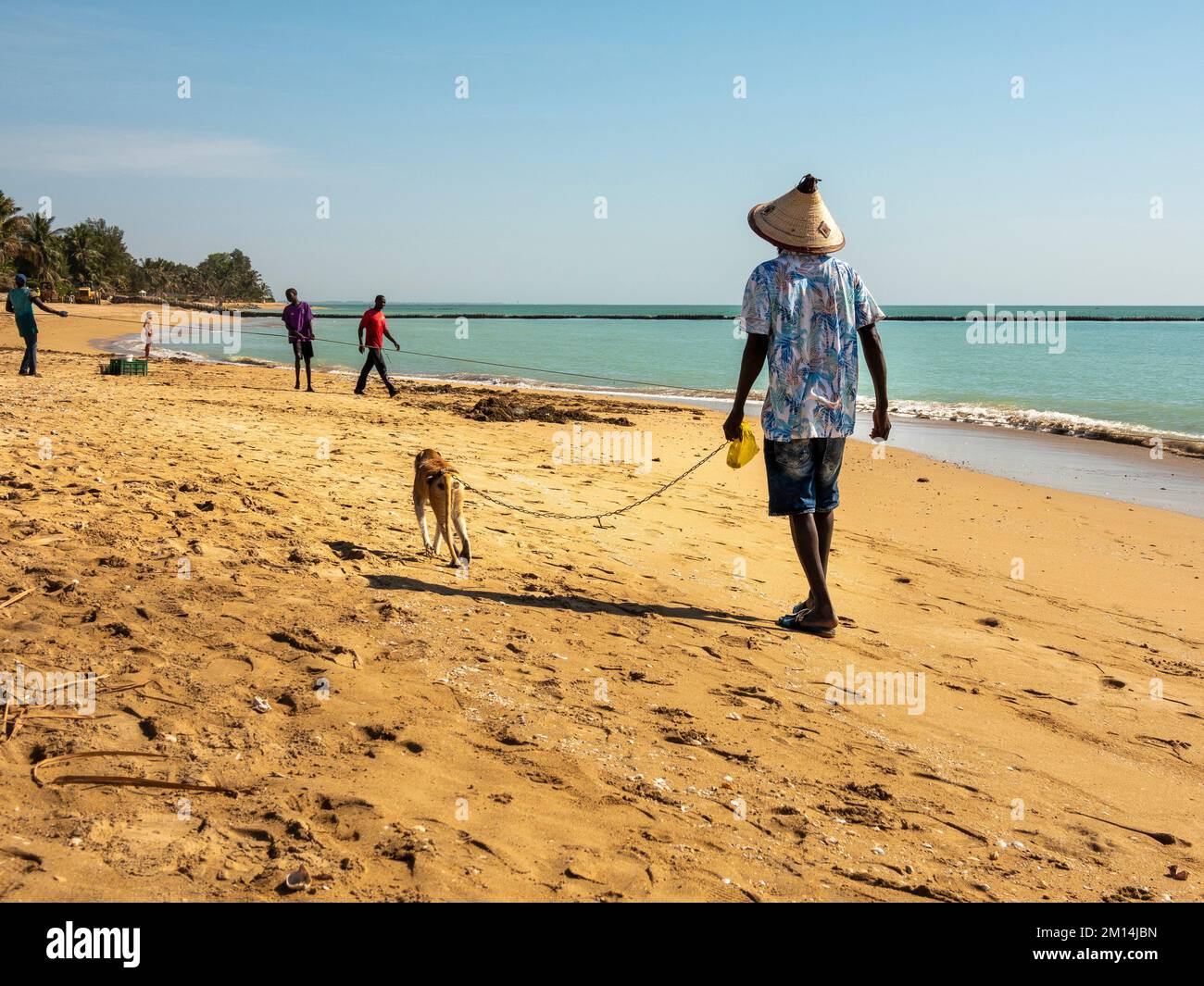 MBOUR, SENEGAL - JANUARY CIRCA, 2022. Scene of unidentified man with hat on beach keeping his monkey on a leash as domestic animal. Monkeys, wild or d Stock Photo