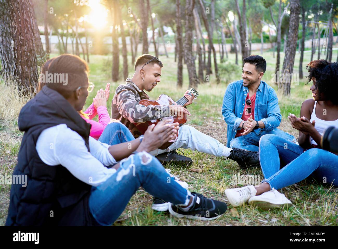 Multi-ethnic friends sitting on the ground while one plays guitar in a ...