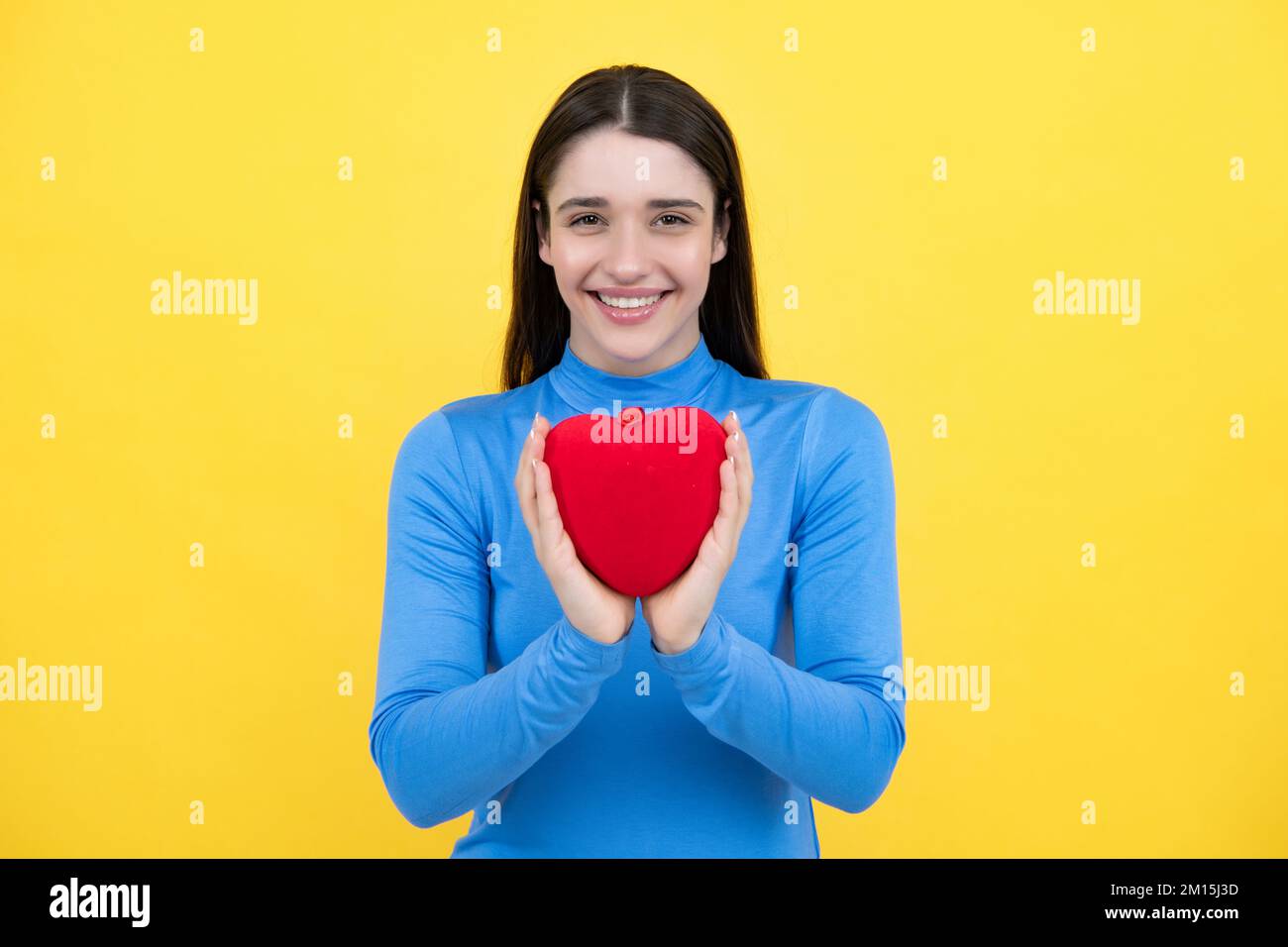 Portrait of lovely sweet cute pretty girl hold red paper card heart isolated over yellow background. Beautiful girl holding heart, love shape. Stock Photo