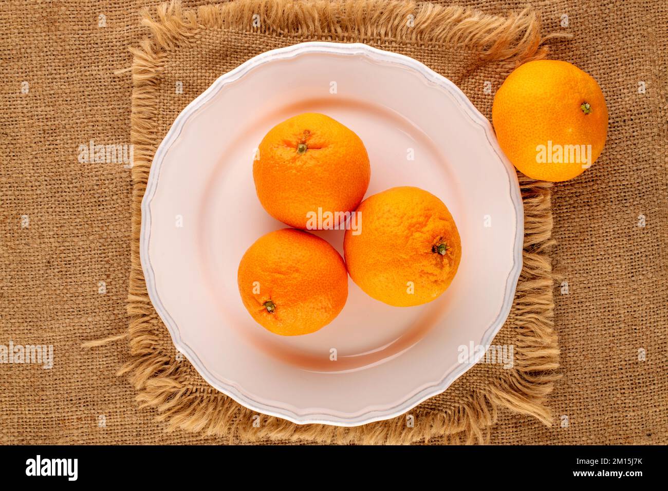 Several ripe tangerines in a white ceramic dish on a jute cloth, macro, top view. Stock Photo