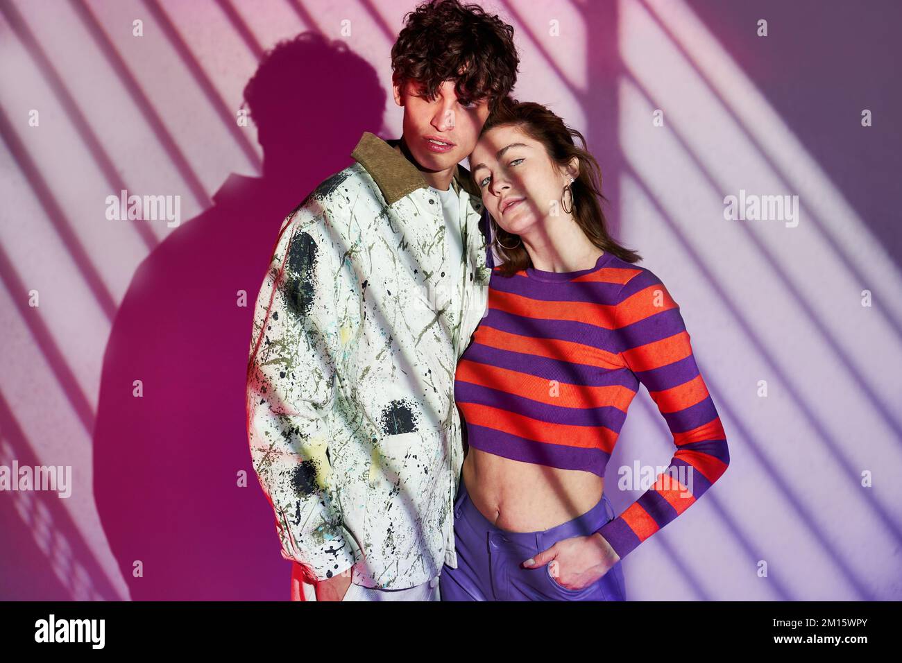 Arrogant boyfriend and girlfriend in colorful street style outfits standing near purple wall and hugging in studio with striped lightning Stock Photo