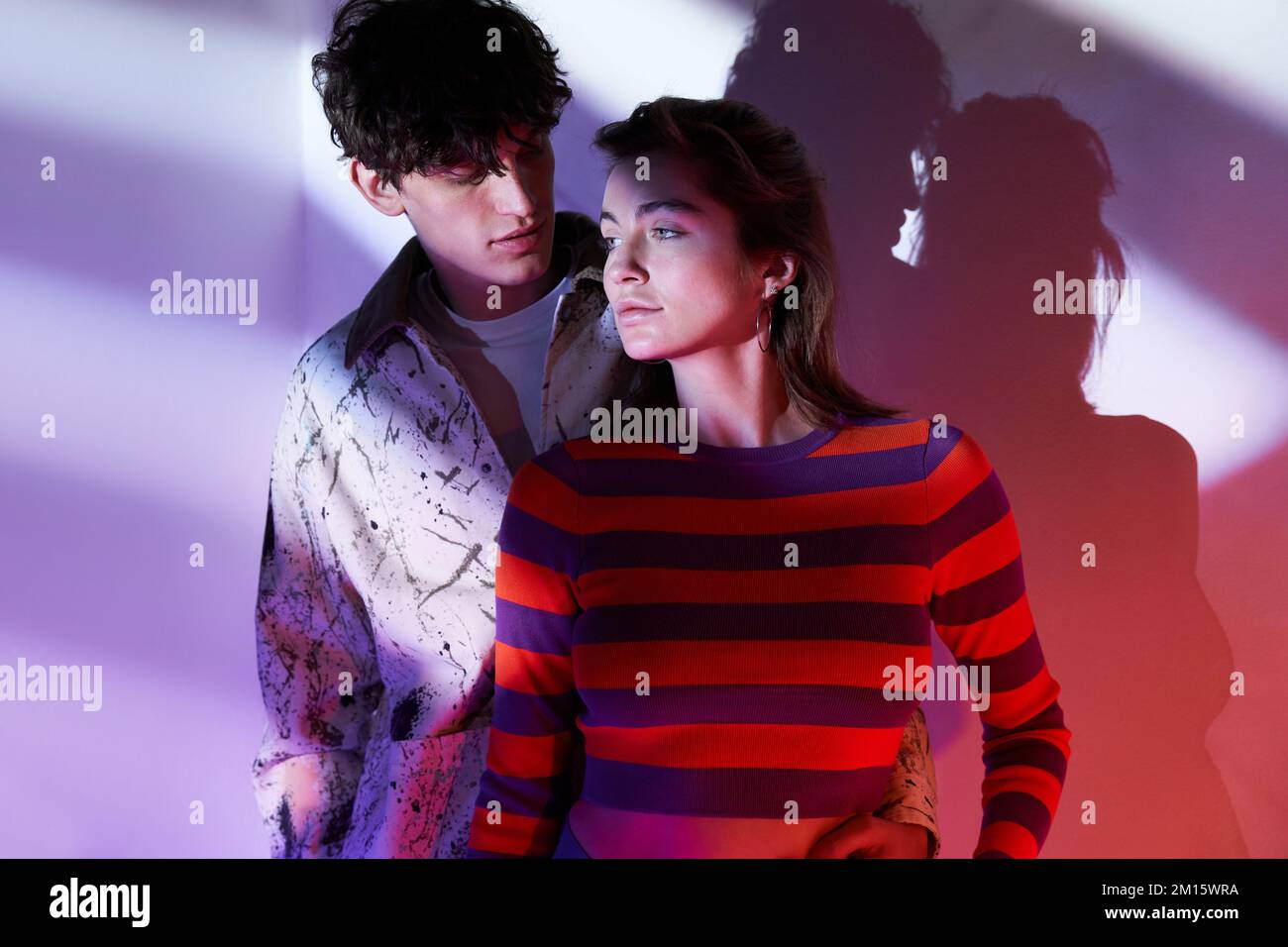 Young models in colorful outfits standing embracing each other near purple wall in neon light in studio Stock Photo