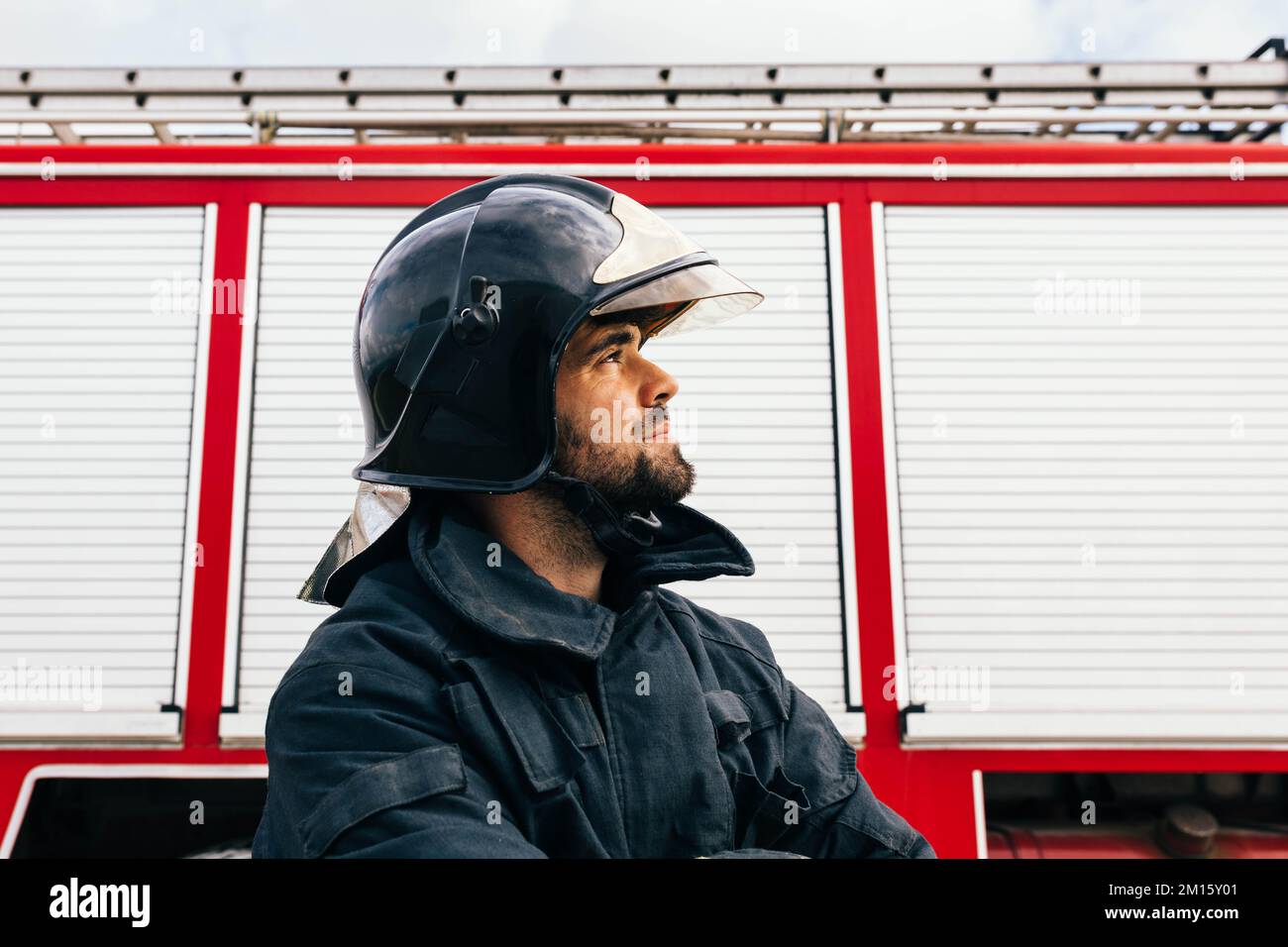 Side view of Hispanic fireman in protective uniform looking away with smile standing near fire engine on station Stock Photo