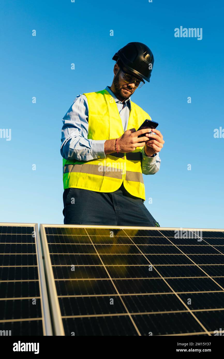 Low angle of positive male engineer in protective helmet and bright waistcoat standing near solar batteries and browsing cellphone on sunny day Stock Photo