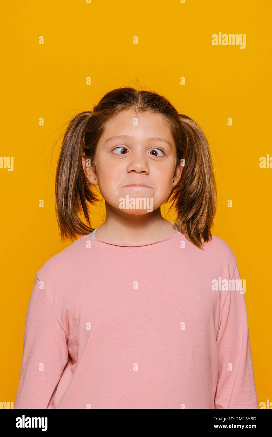 Young little girl in casual outfit looking at camera with astonished face expression. She is going cross-eyed while standing on yellow background in s Stock Photo