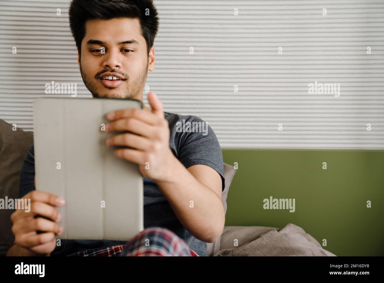 Young handsome smiling indian man holding tablet looking on it while sitting at home Stock Photo