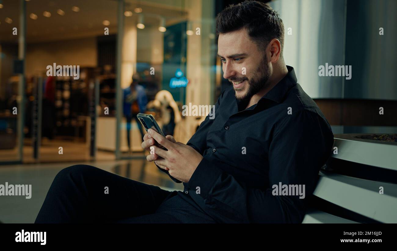 Caucasian stylish man in black suit successful businessman customer sit at shopping mall using mobile phone apps make online order buying clothes win Stock Photo