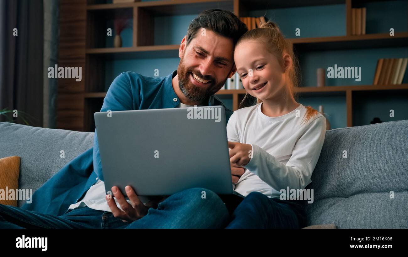 Cheerful father and little beautiful daughter girl sit on couch watch movies cartoons on internet spend time together at home having fun use laptop Stock Photo