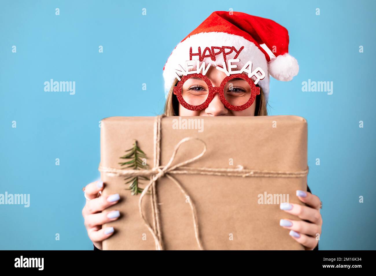 Beautiful, young, blonde girl with Santa hat and prop glasses isolated on blue background, peaking behind a gift box. Copy space Stock Photo