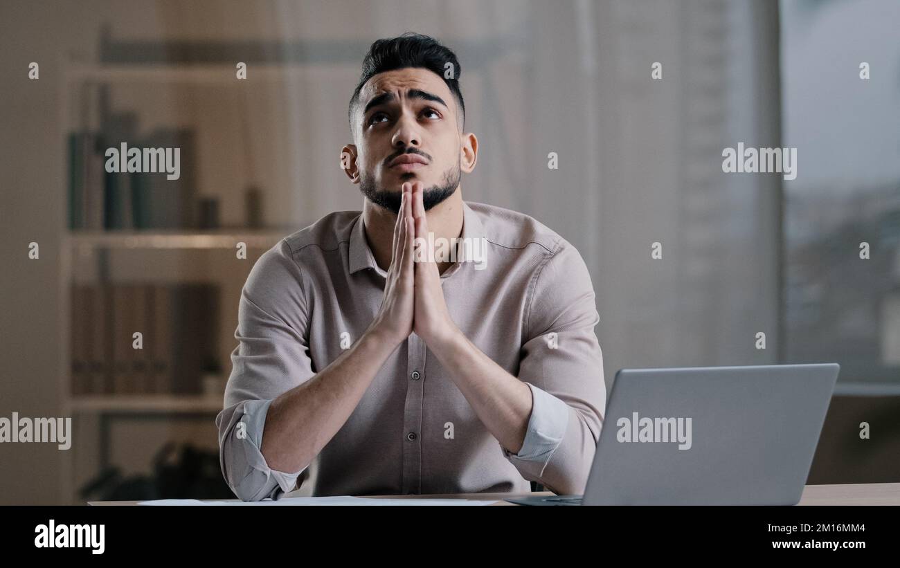 Frustrated young hispanic male analyst religious business man feel stress check financial documents ask for help concentrate mind at problem sit with Stock Photo