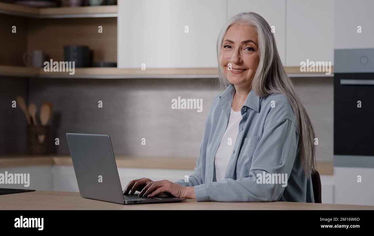 Caucasian senior teacher elderly housewife businesswoman grandmother grey-haired lady work on laptop in kitchen use home wifi booking tickets chatting Stock Photo
