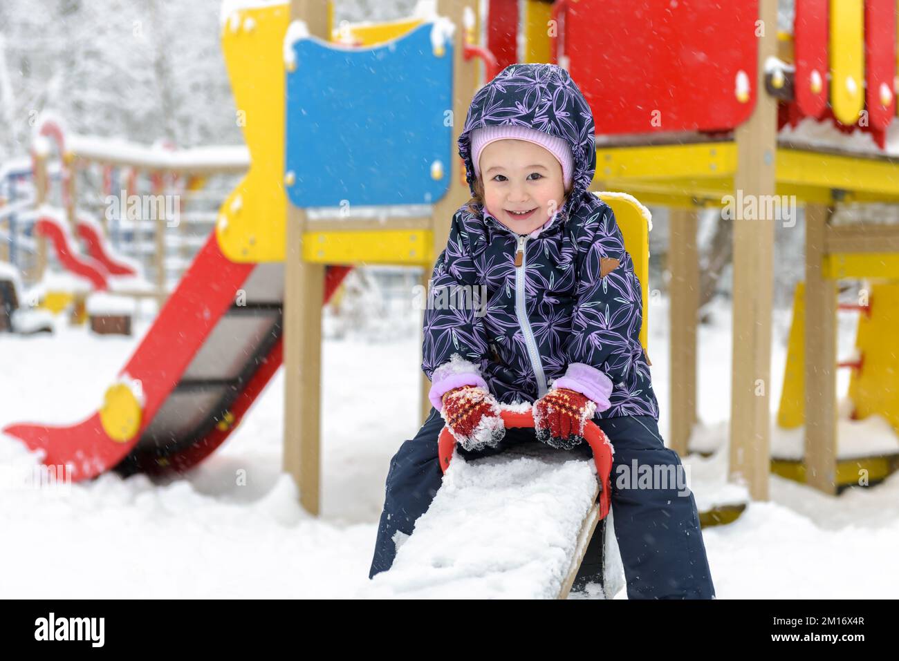 Child on seesaw in winter, little kid having fun on snowy playground. Baby girl plays and looks at camera in urban park. Theme of snow, game, Christma Stock Photo