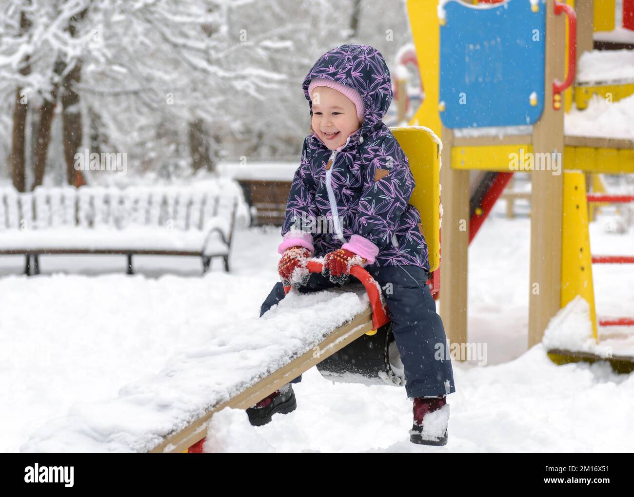 Child having fun on seesaw in winter, happy baby girl playing in snowy park. Little kid is on playground outdoor. Theme of snow, winter, game, cold an Stock Photo