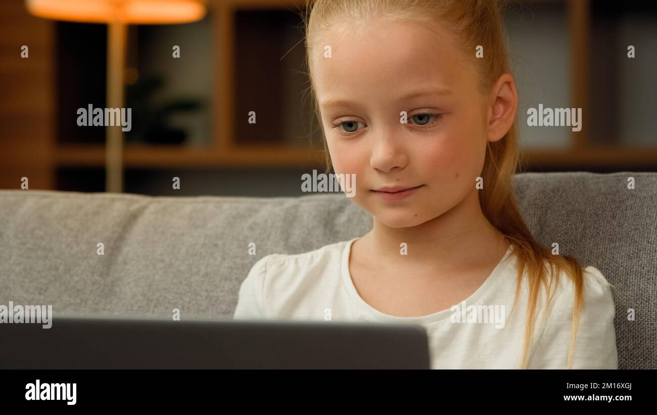 Adorable preteen little girl sitting on couch using modern laptop to watch cartoon movie funny video looking at computer screen laughing studying onli Stock Photo