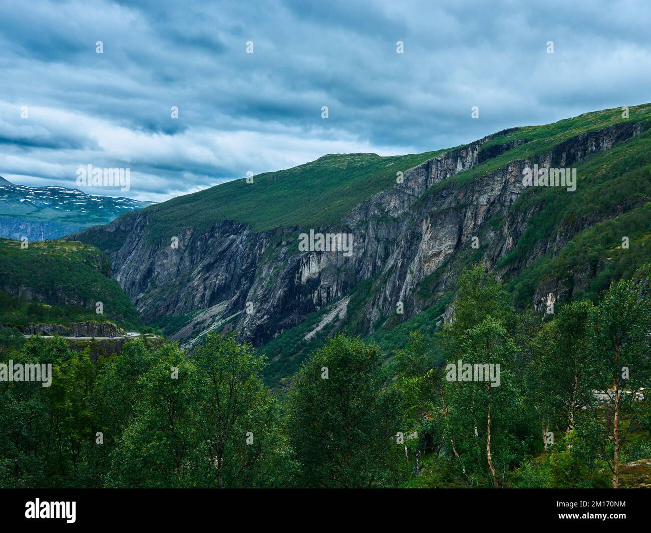 A rocky mountain with slopes covered with grass, surrounded by green trees, with cloudy sky in the background Stock Photo