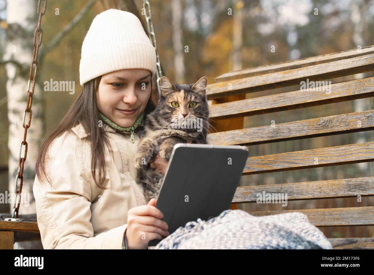 Girl uses a tablet while sitting on a bench in an autumn park with a cat in her arms. Stock Photo