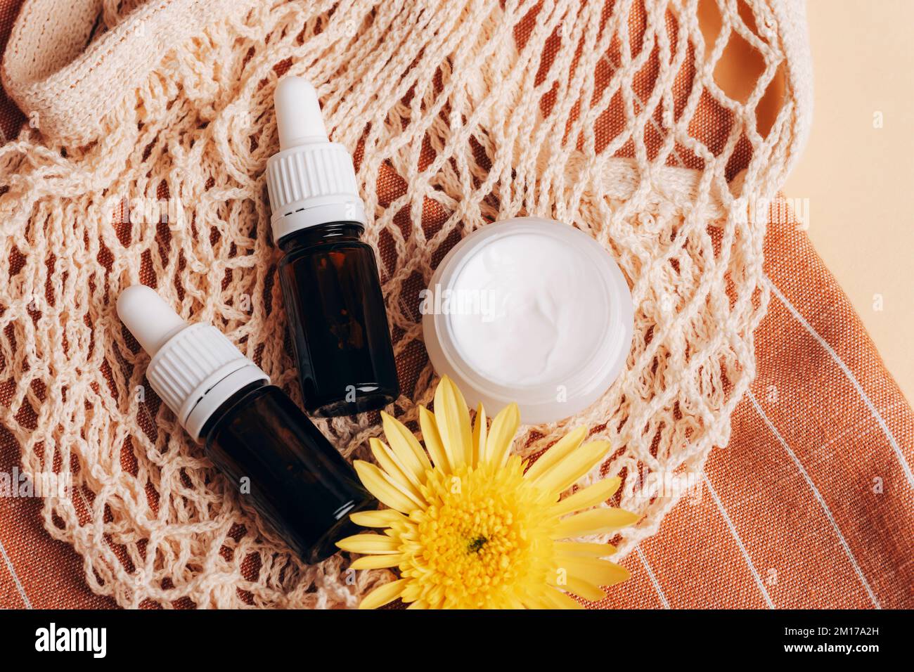 Cosmetic bottles and cream jar with yellow chrysanthemum flower on mesh bag. Organic spa cosmetic beauty product. Zero waste concept. Stock Photo