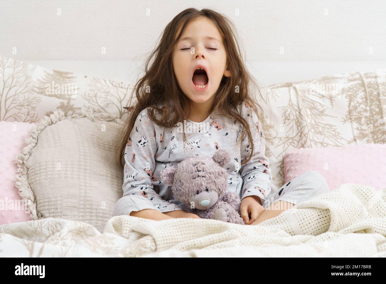 Funny, cute, tired little brunette girl yawning with open mouth, carrying teddy bear toy, sit on soft bed with pillows on white background. Broken sle Stock Photo