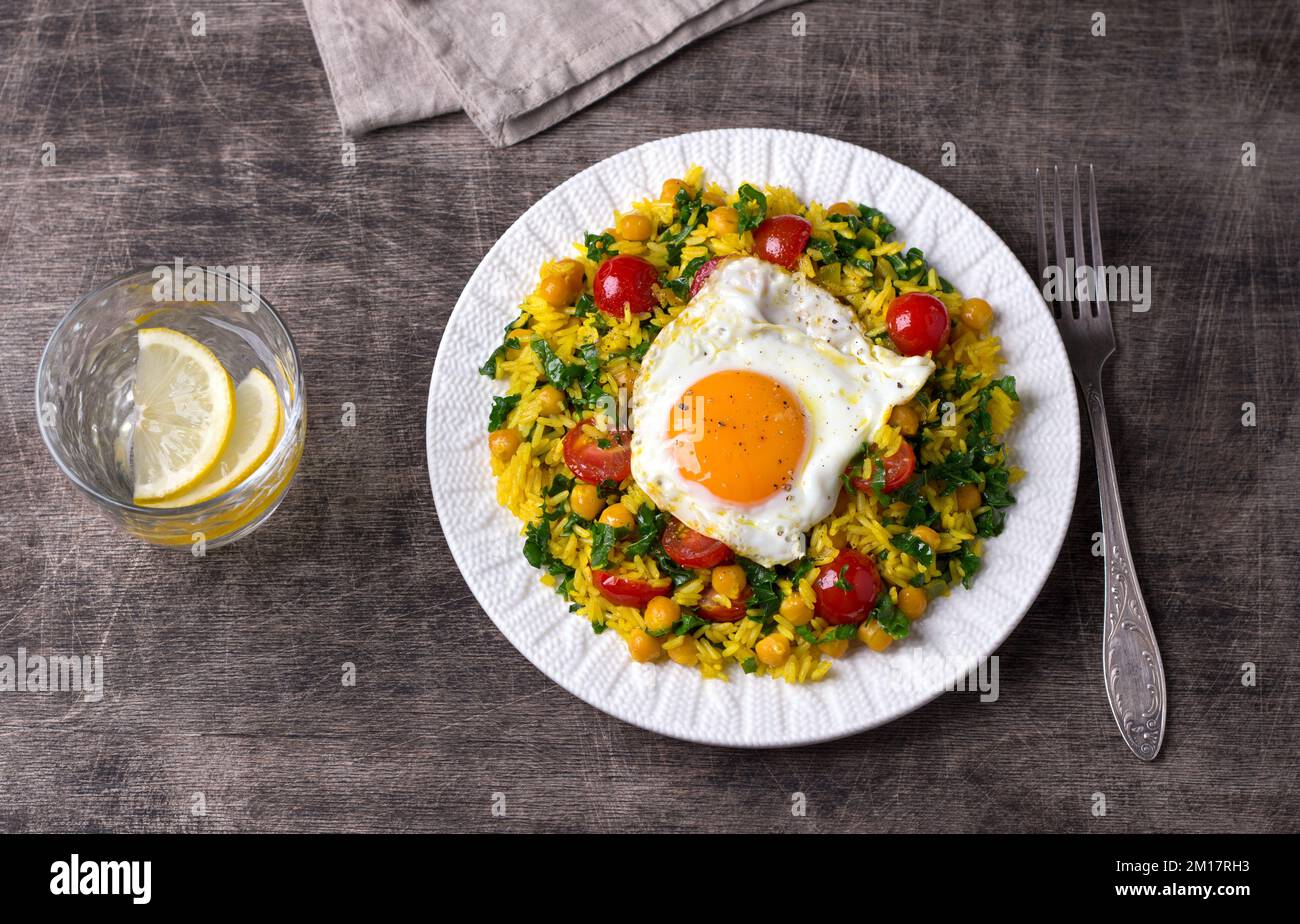 Chinese fried rice with turmeric, chickpeas, tomatoes, kale and with fried eggs on white ceramic plate on dark wooden background. Quick and easy dinne Stock Photo